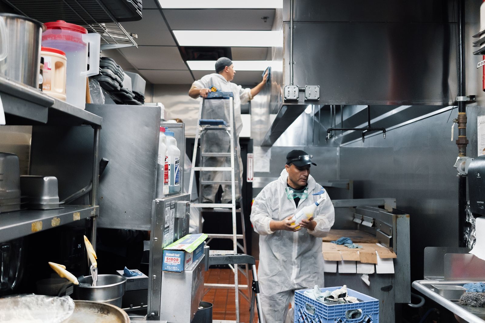 Two TruShine crew members cleaning commercial kitchen hood system with one on ladder wiping exterior and one cleaning below
